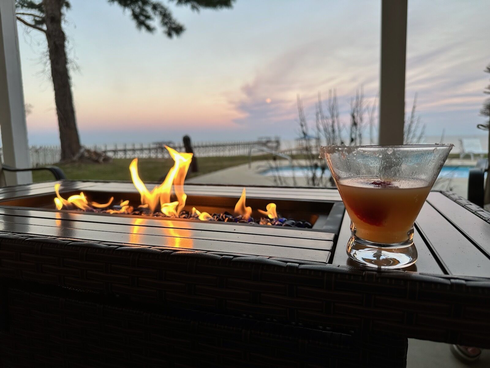 A glass of drink on a table with a fire feature in the background.