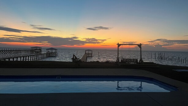 Un espace piscine offrant une vue sur le coucher de soleil sur l’océan, une jetée s’avançant dans l’eau et un kiosque.