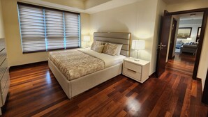 A bedroom with a large bed, bedside tables with lamps, a window with blinds, and a wooden floor. - Opulent Comfort Suites  (Colombo)