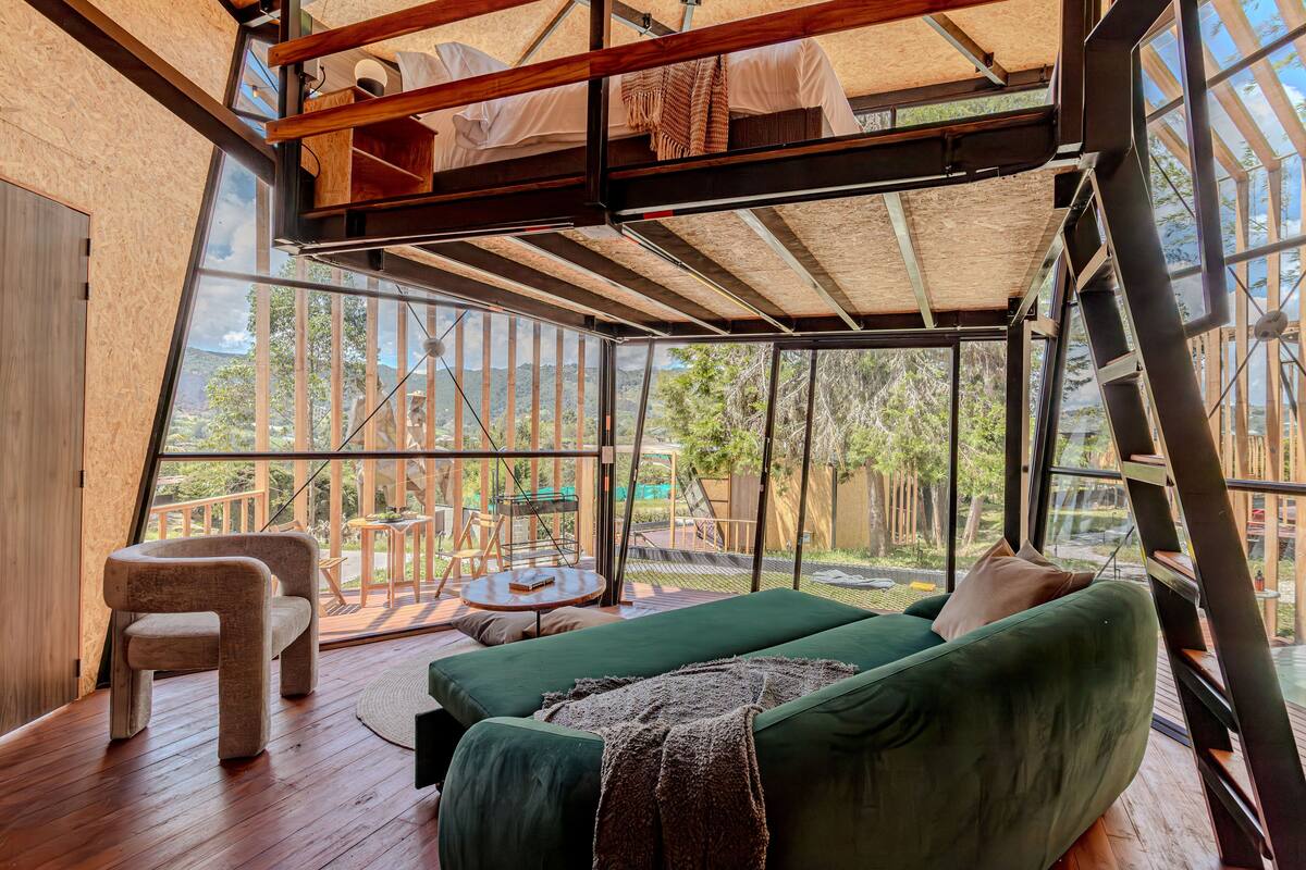Una habitación amplia con un sofá verde, una silla de madera y una pequeña mesa redonda. Se ve una cama litera con ropa de cama blanca al fondo.