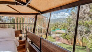 Un dormitorio con cabecera de madera, cama con ropa de cama blanca y un ventanal con vistas a un jardín y montañas.