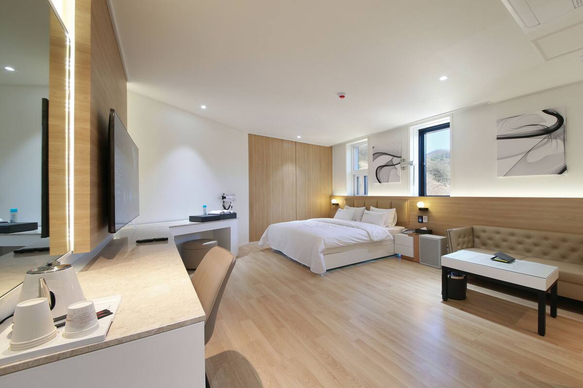 A modern hotel room with a bed, bedside table, a desk with a chair, a sofa, and artwork on the walls.