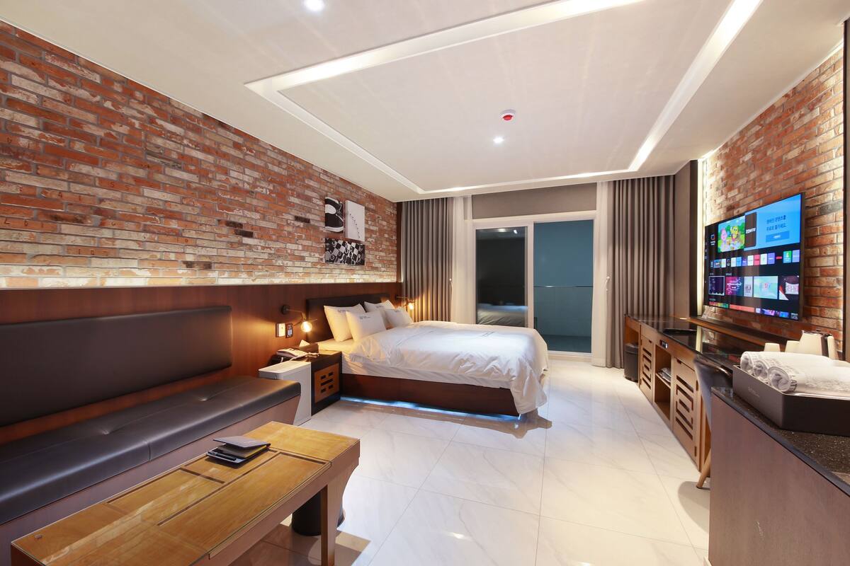 A modern hotel room with a brick accent wall, a large bed, a flat-screen TV, a wooden bench, and a desk with a lamp.