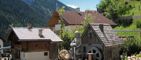 A rustic building with a water feature, surrounded by greenery and mountains in the background.