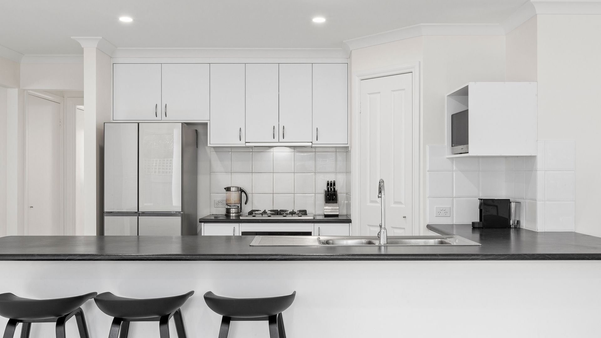 A modern kitchen with white cabinetry, a stainless steel refrigerator, a gas stove, and a sink. There are three black bar stools in front of the counter.