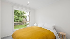 Ein Schlafzimmer mit einem großen Fenster, einem Bett mit weißer Bettwäsche und einem kleinen Holzschemel.