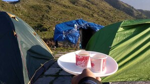 A person holding a tray with two cups of coffee, a tent, and a stone pathway. - Triund Trek and Camping (Dharamshala)