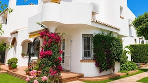 Un bâtiment blanc à la façade incurvée, un balcon orné de fleurs roses et un chemin de briques menant à l’entrée.