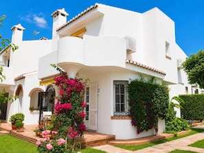 Un bâtiment blanc à la façade incurvée, un balcon orné de fleurs roses et un chemin de briques menant à l’entrée.