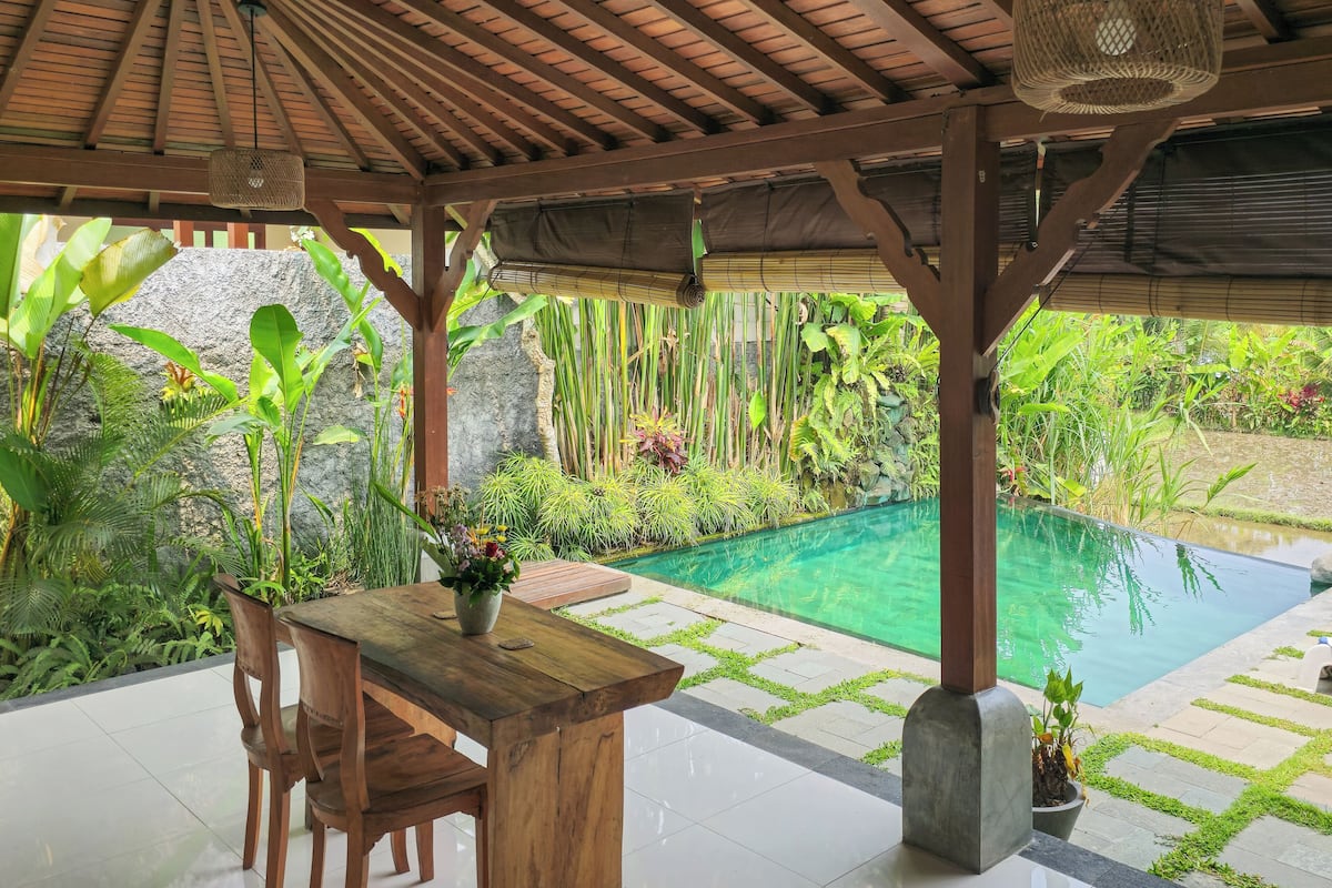 A wooden table and chairs under a thatched roof, with a pool and lush greenery in the background.