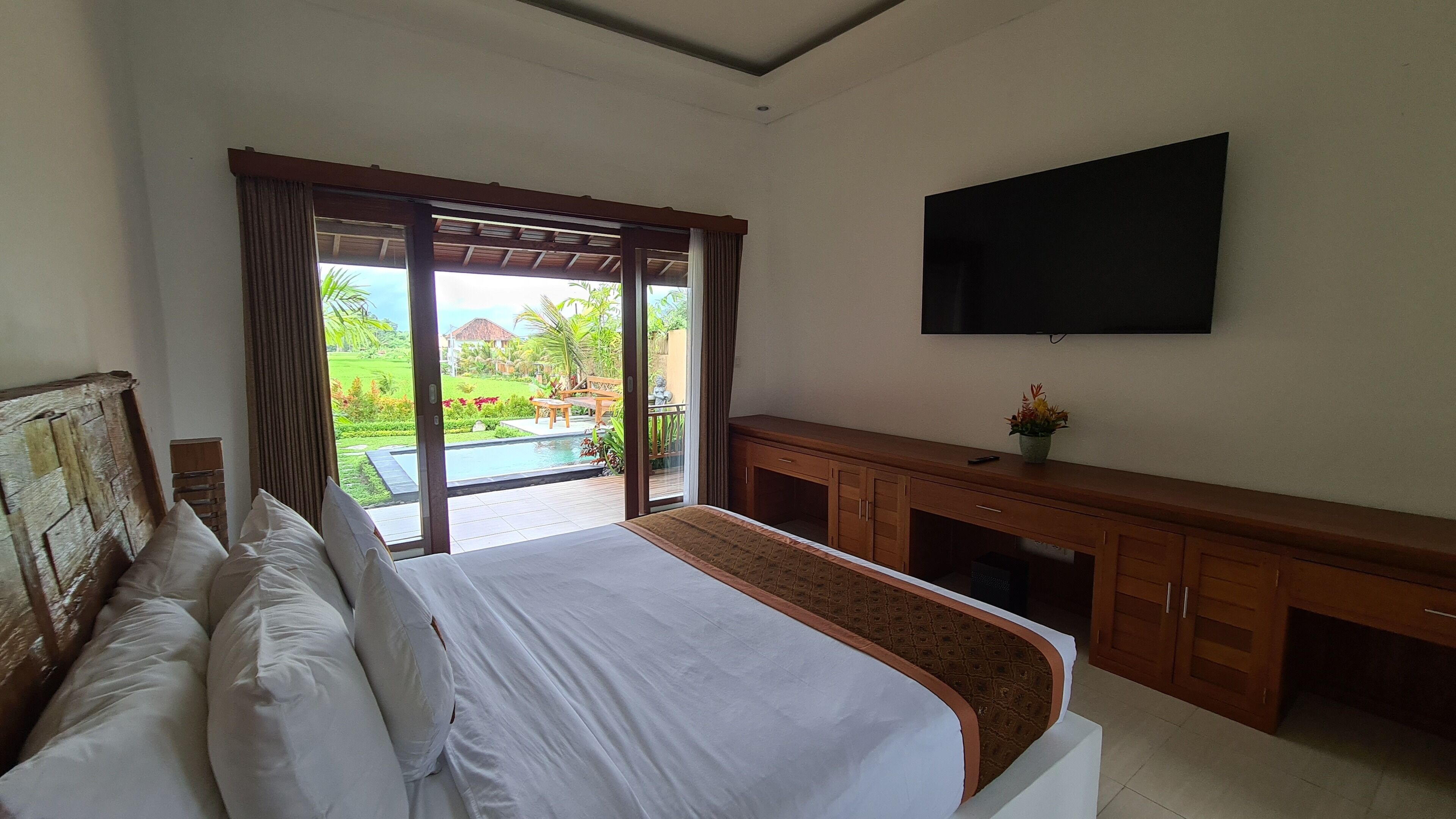 A bedroom with a large bed, a TV mounted on the wall, and a view of a pool and garden outside.