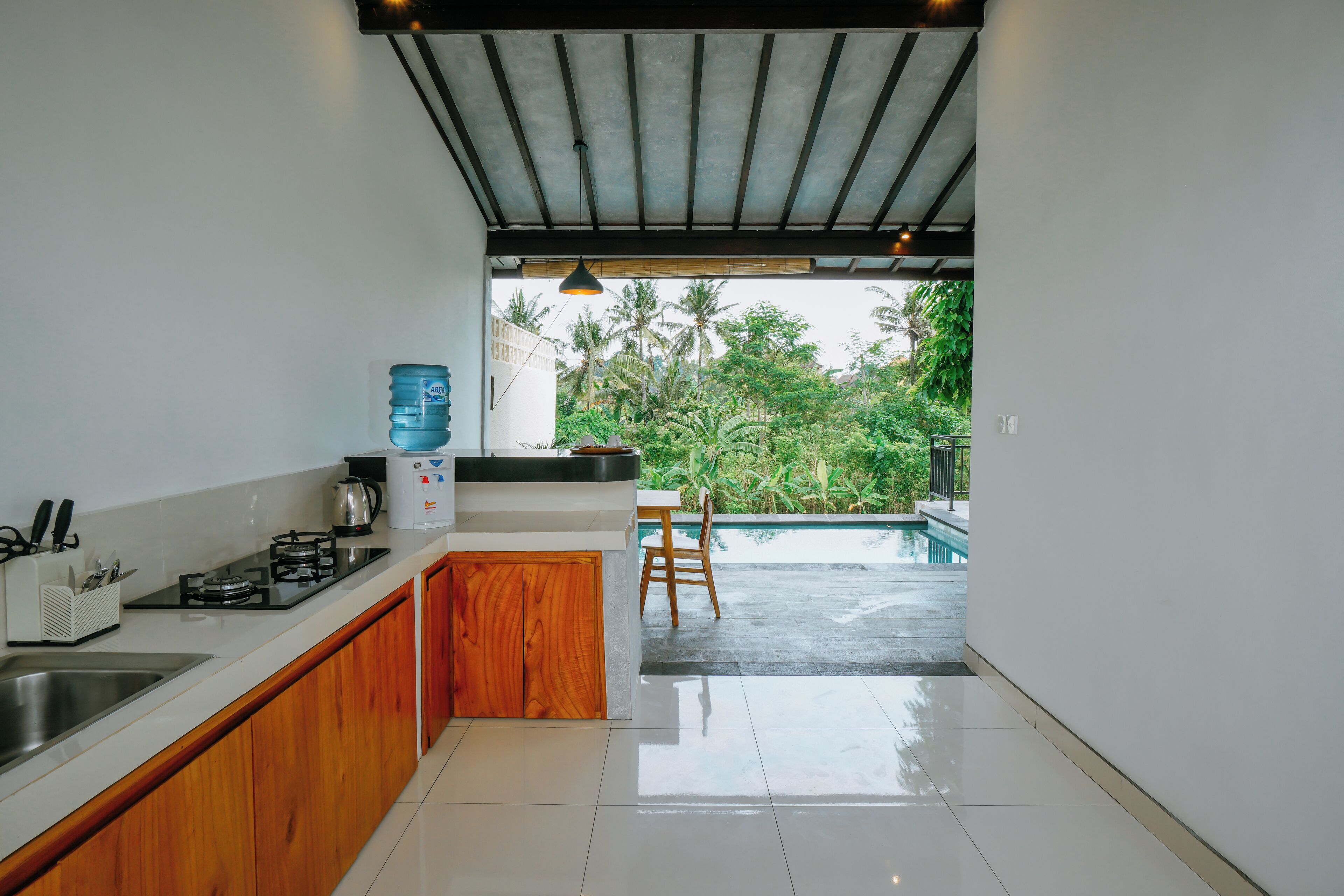 A modern kitchen with a gas stove, sink, and wooden cabinets. There is a view of a swimming pool and lush greenery outside.