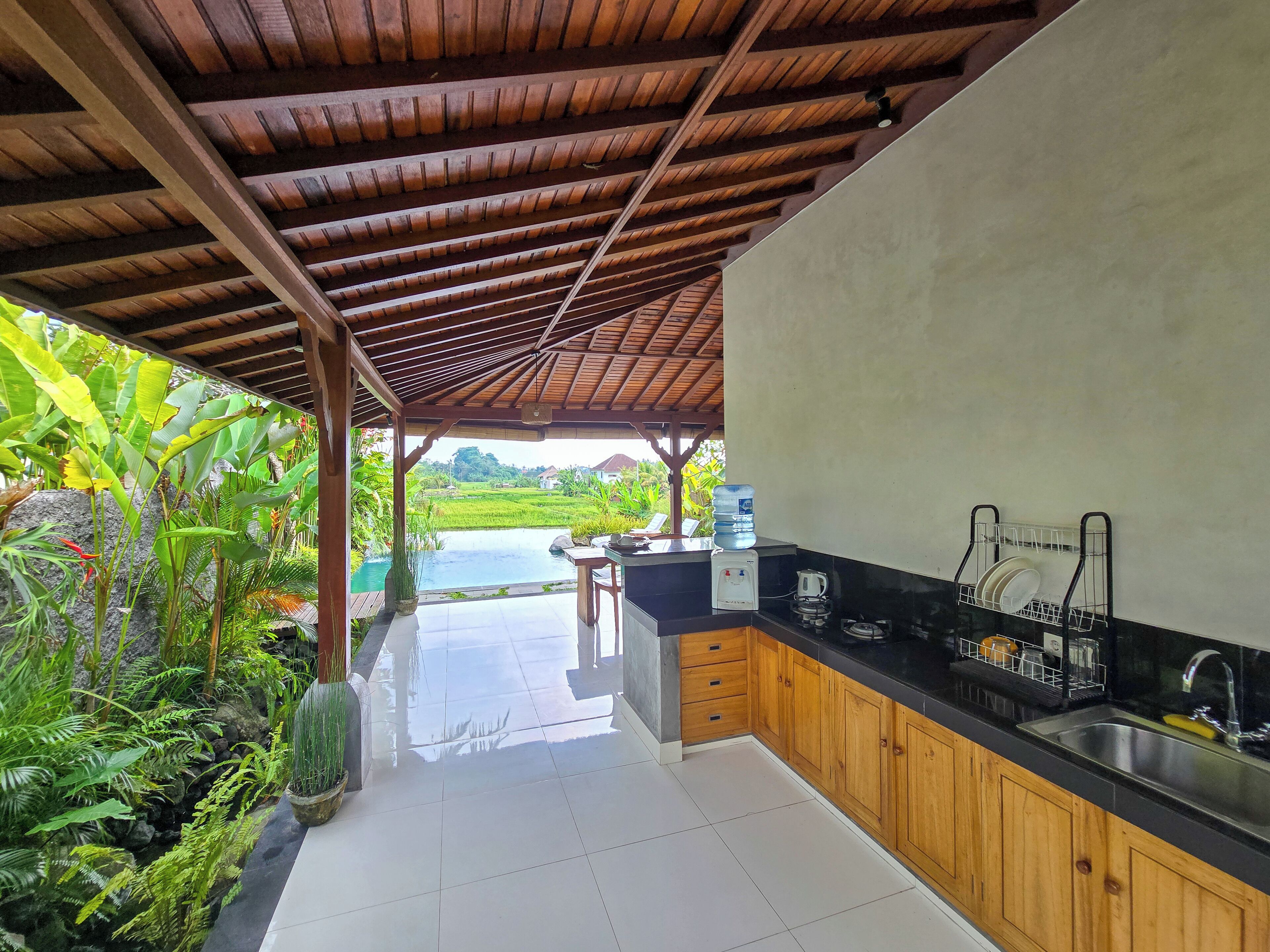 A kitchen with a wooden countertop, a sink, and a water dispenser. There is a view of a swimming pool and lush greenery outside.