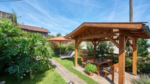 Eine hölzerne Pergola mit Tisch und Stühlen, umgeben von üppigem Grün und blauem Himmel.