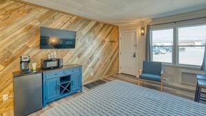 A modern hotel room with a herringbone wood wall, a flat-screen TV, a kitchenette with a coffee maker, a blue chair, and a large window offering a view of the parking lot. - Comfy and Cozy Studio for 2 (101 Spring Creek) (Kalispell)