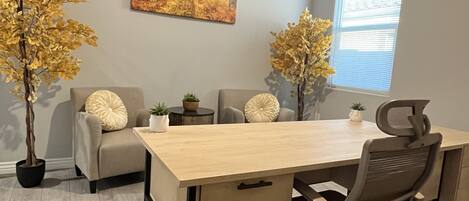 A modern office space with a wooden desk, two grey chairs, a sofa, and two potted plants.