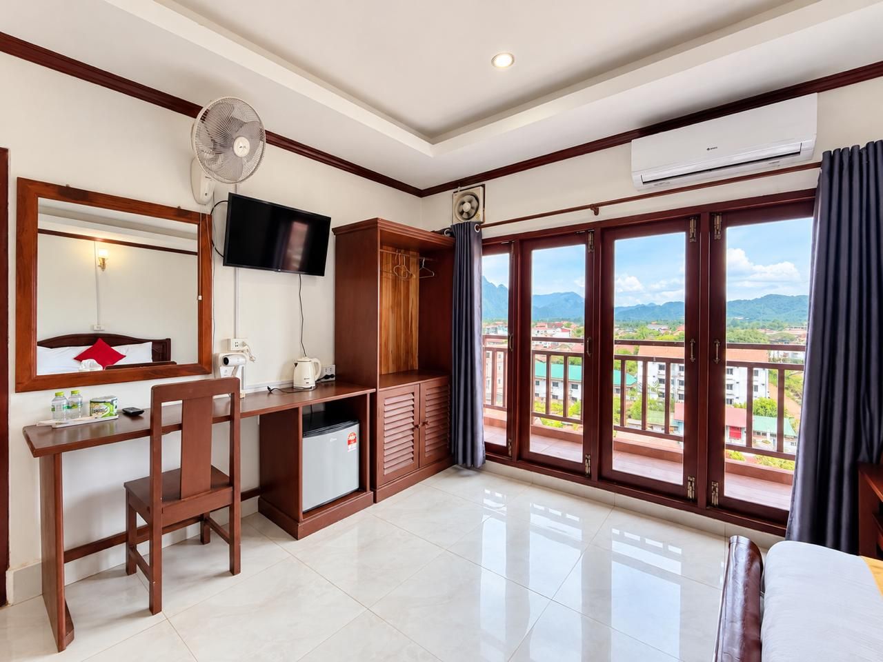A hotel room with a desk, chair, TV, wardrobe, and a balcony with a view.