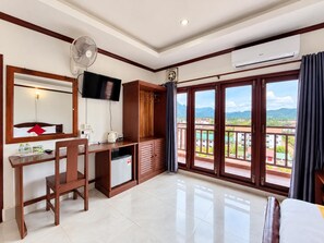 A hotel room with a desk, chair, TV, wardrobe, and a balcony with a view.