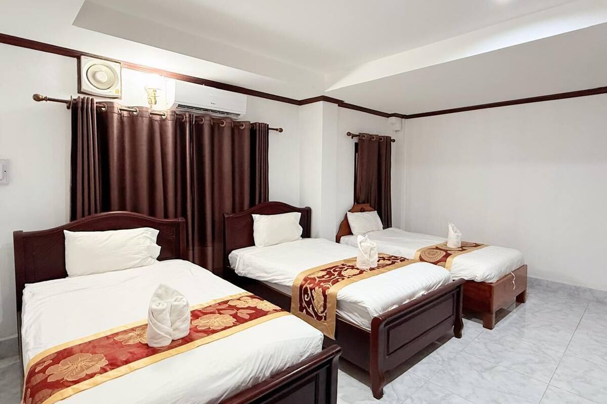 A hotel room with three wooden beds, each with white linens and a red and gold decorative headboard. There is a ceiling fan, a wall-mounted air conditioner, and a window with dark curtains.