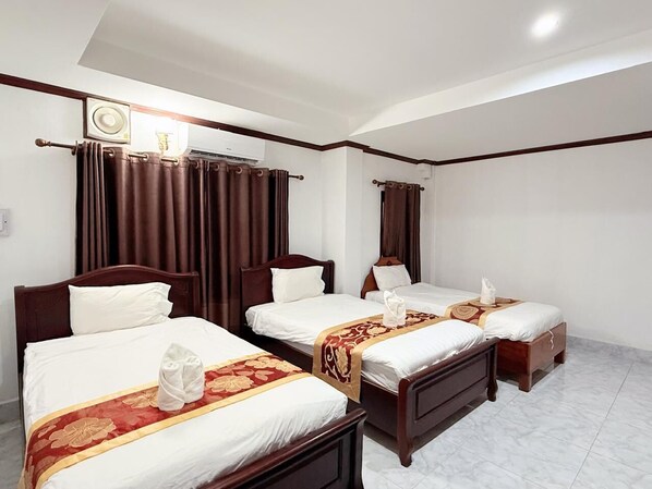 A hotel room with three wooden beds, each with white linens and a red and gold decorative headboard. There is a ceiling fan, a wall-mounted air conditioner, and a window with dark curtains.