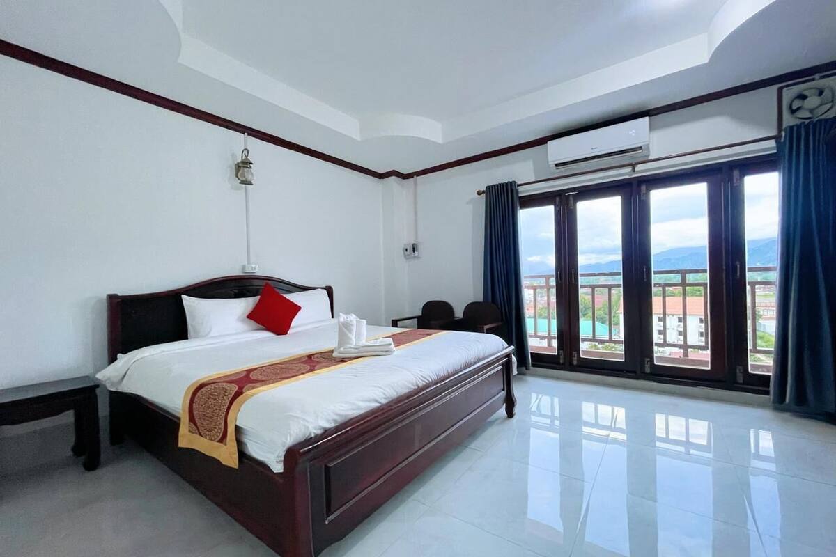 A hotel room with a large bed, a desk, and a view of the outdoors through large windows.