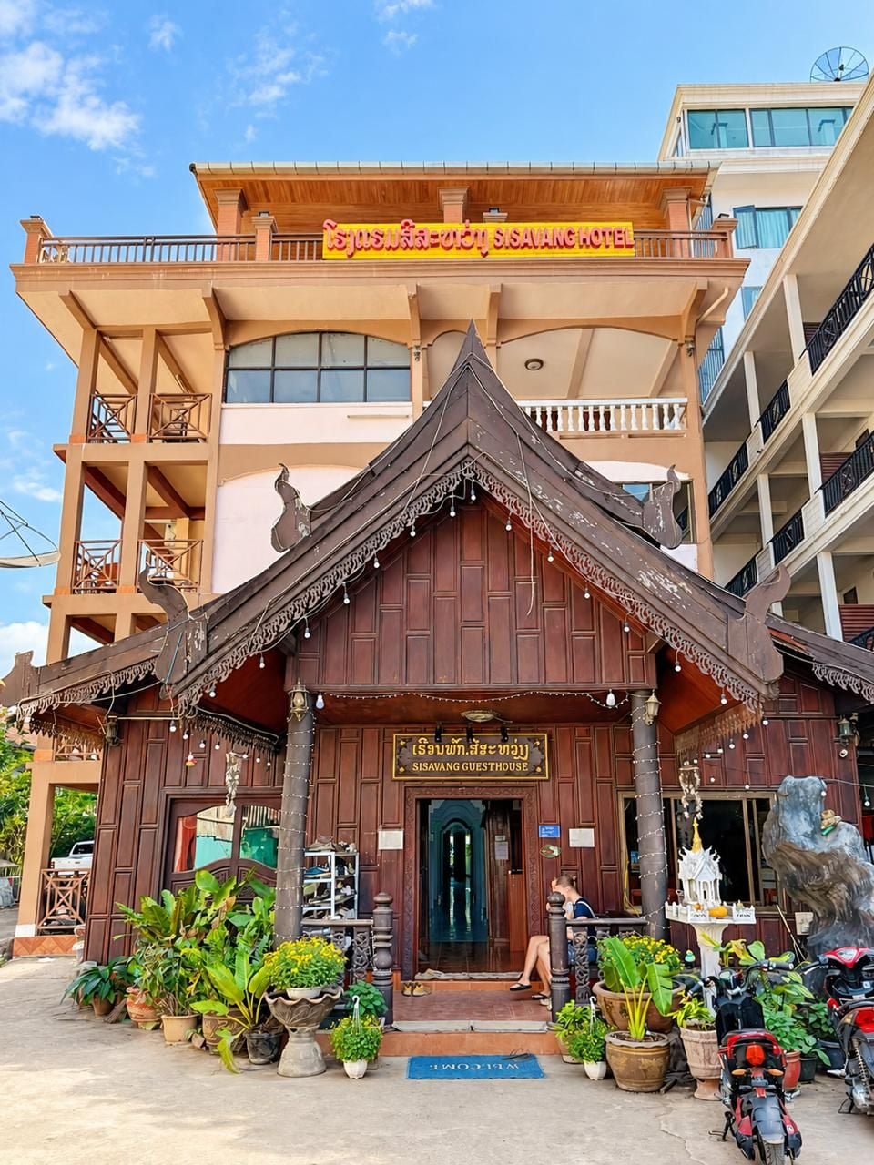 A traditional wooden guesthouse with a signboard, potted plants, and a motorcycle parked in front.