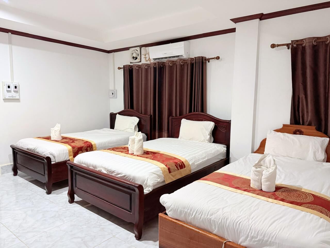 A hotel room with four beds, each with a wooden frame and white bedding. There are brown curtains on the windows, and an air conditioning unit is mounted on the wall.