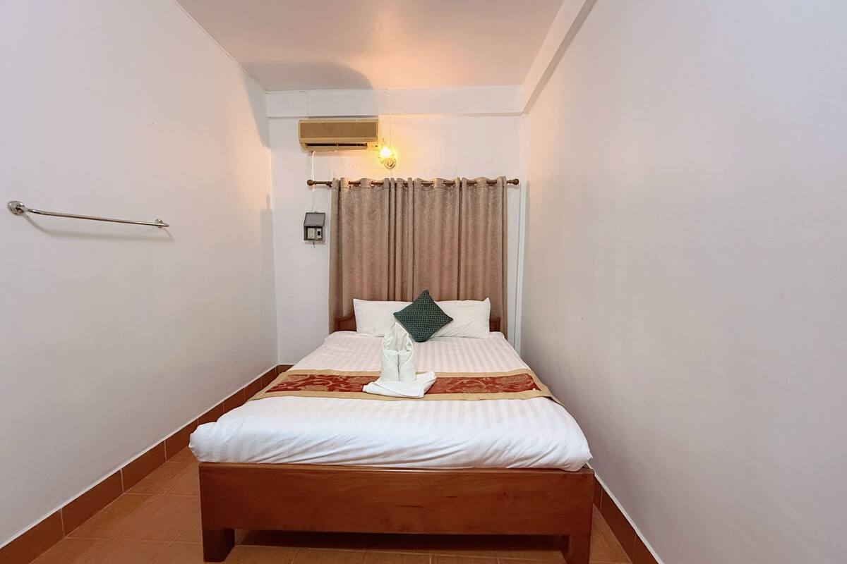 A neatly made bed with white bedding and a brown headboard, a bedside table with a lamp, and a wall-mounted air conditioning unit.