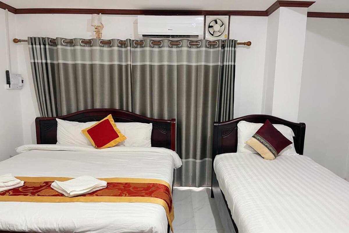 A hotel room with two single beds, a large window with curtains, an air conditioning unit, and a wall-mounted clock.