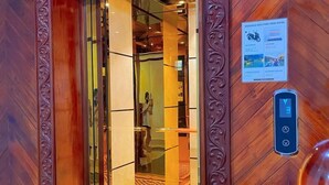 Un ascenseur aux panneaux de bois richement décorés et doté d’une porte en verre, qui donne sur une pièce au sol à carreaux et où l’on aperçoit un lit.