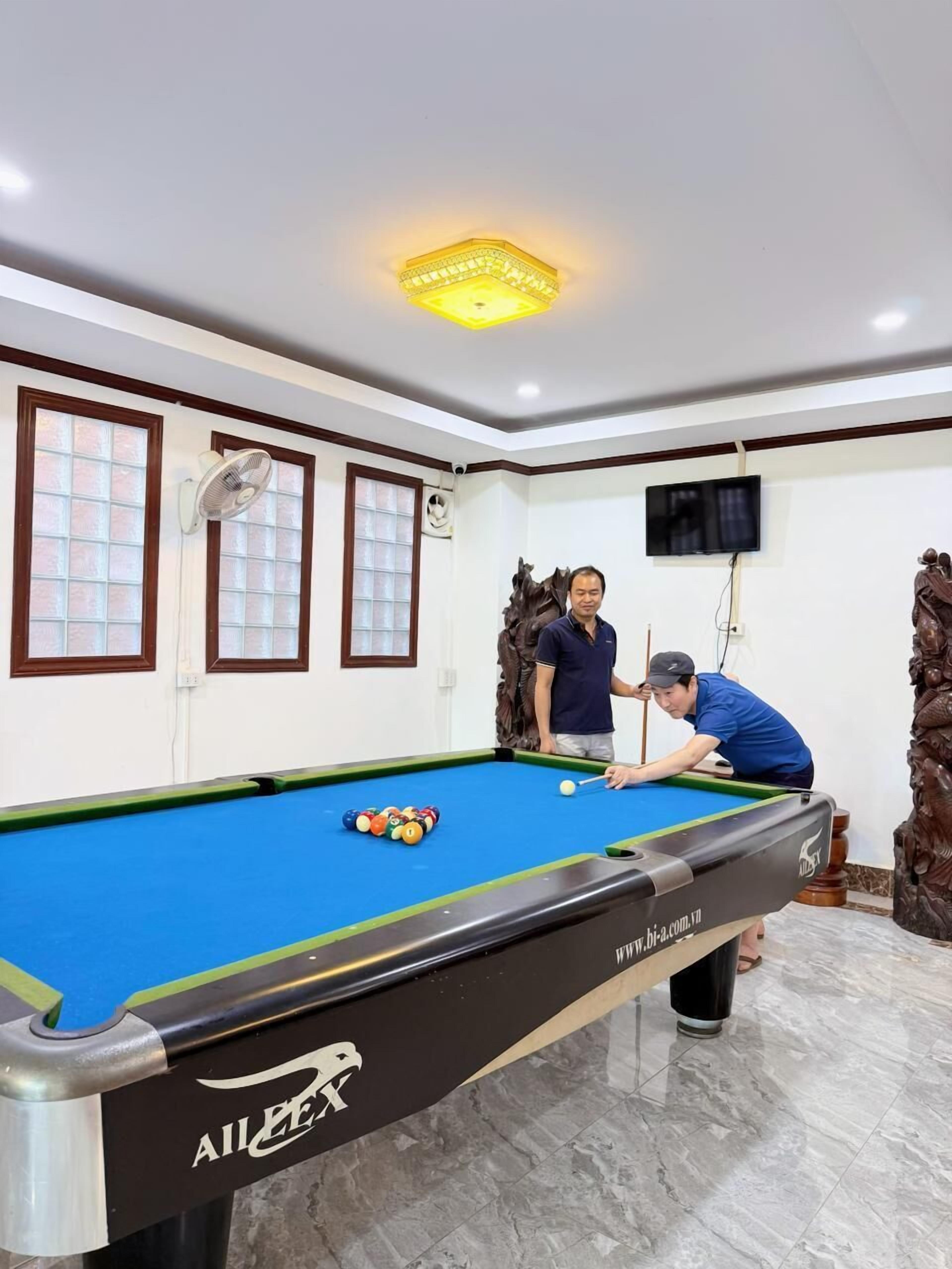 A pool table with two people playing, a television mounted on the wall, and a decorative wall piece.