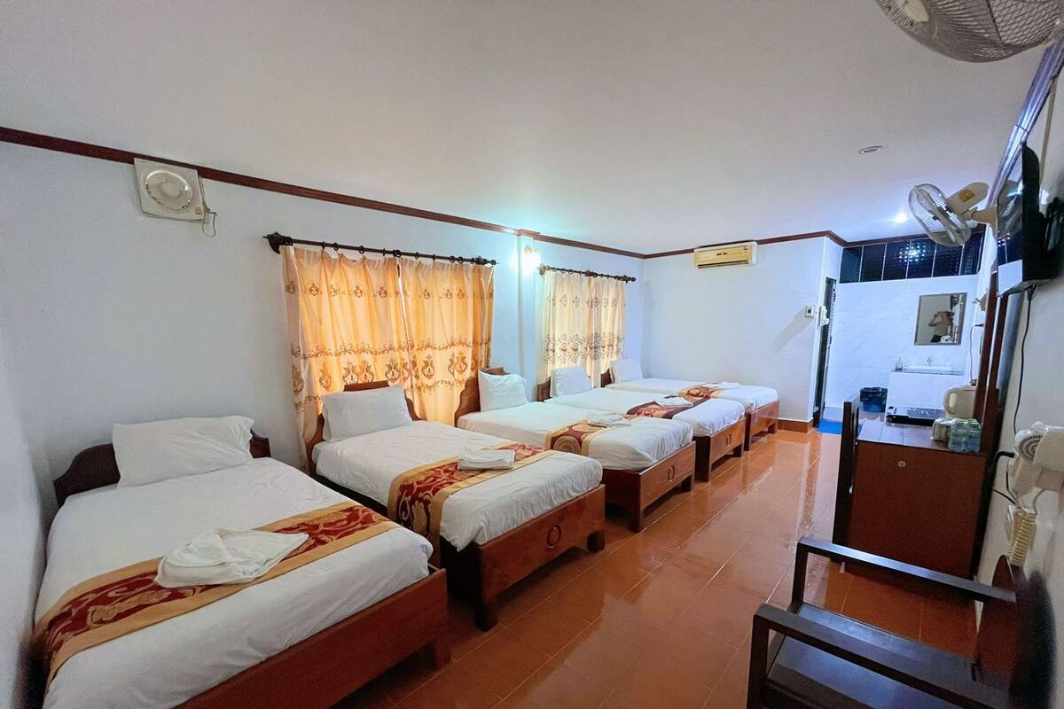 A hotel room with four single beds, a wooden dresser, a desk with a chair, and a ceiling fan.