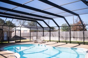 A covered backyard with a swimming pool, lounge chairs, and a wooden fence.
