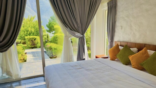 A bedroom with a large bed, grey curtains, and a view of greenery outside.