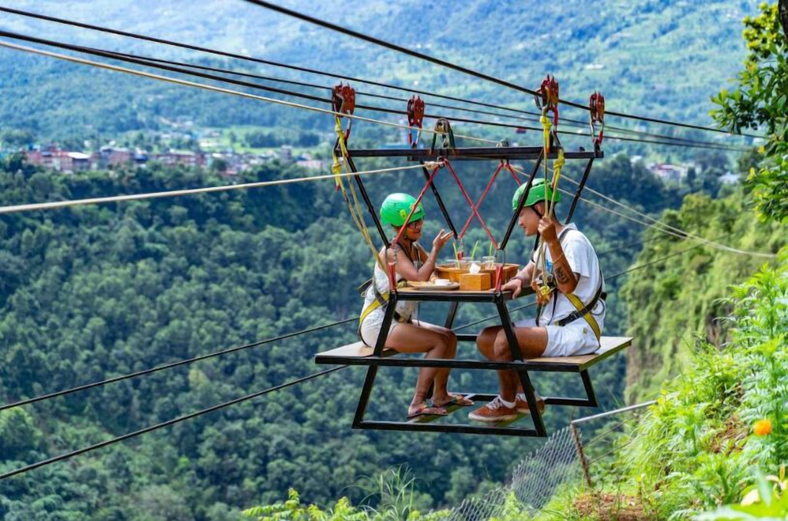 Two people on a suspended platform with a table, overlooking a forested valley.