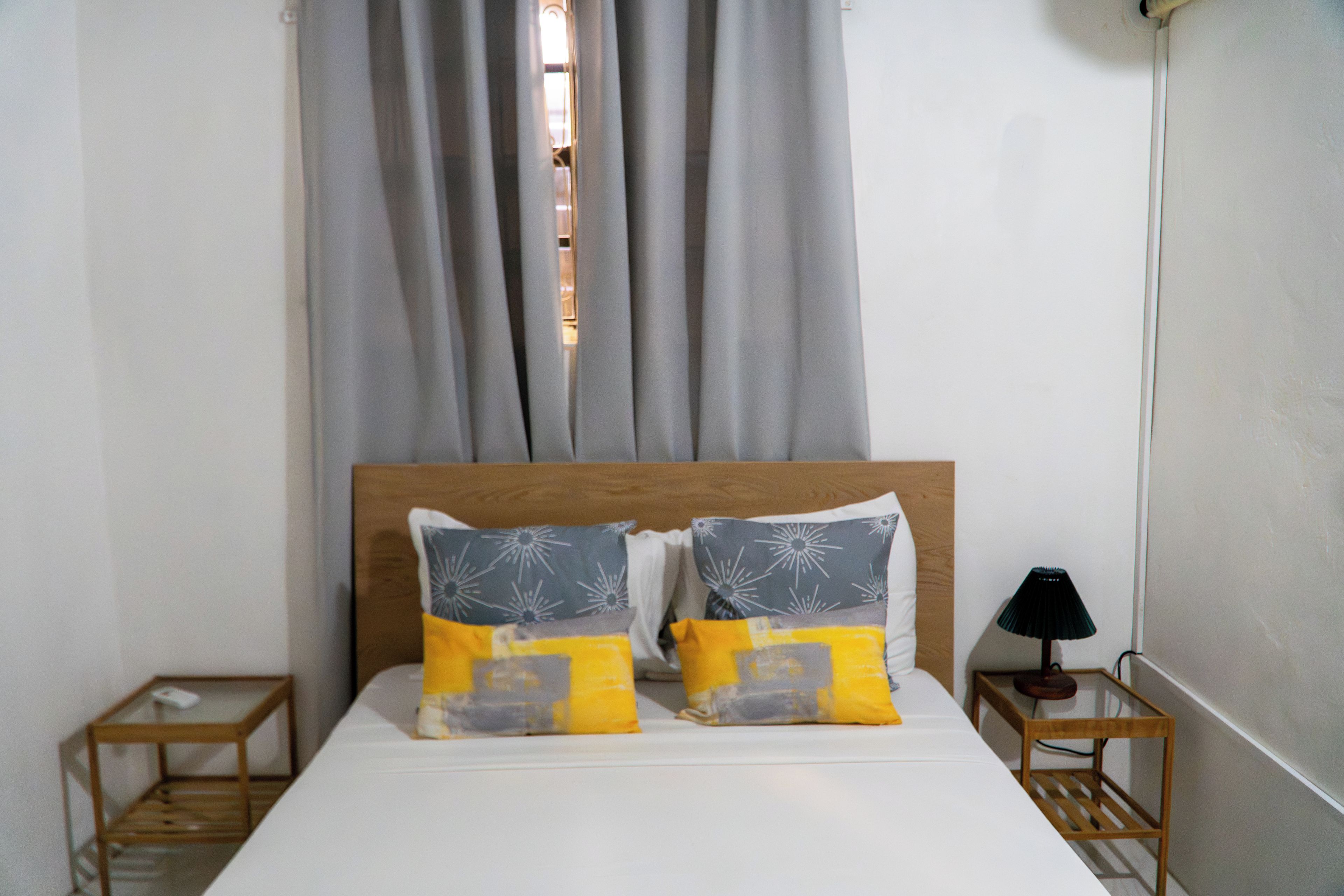 A bed with two pillows, a wooden headboard, a nightstand with a lamp, and a window with curtains.