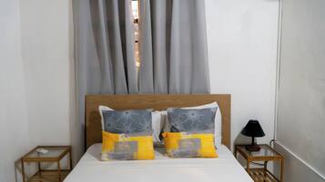 A bed with two pillows, a wooden headboard, a nightstand with a lamp, and a window with curtains.