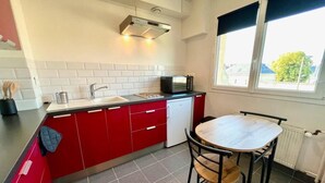 A compact kitchen with a sink, microwave, and red cabinets. There's a window providing natural light and a view of the outside. - Studio Tout Confort Giena-sils (Caen)
