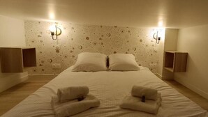 A hotel room with a large bed, two wall-mounted lamps, and a wall clock. - Appartement Antarès-sils (CAEN)