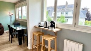 A dining area with a table and chairs, a radiator, and large windows with a view of houses. - Studio Lumineux Khara-sils (Caen)