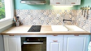 A compact kitchen with a window, a built-in oven, white cabinets, and patterned backsplash tiles. - Studio Lumineux Khara-sils (Caen)
