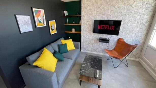 A living room with a grey sofa, yellow and teal cushions, a black coffee table, and a mounted TV displaying "NETFLIX". - Grand Logement Jupiter-sils (CAEN)