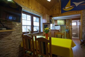 Interior em madeira com área de jantar, mesa e cadeiras amarelas, cozinha com armários de madeira e uma grande janela que oferece luz natural.