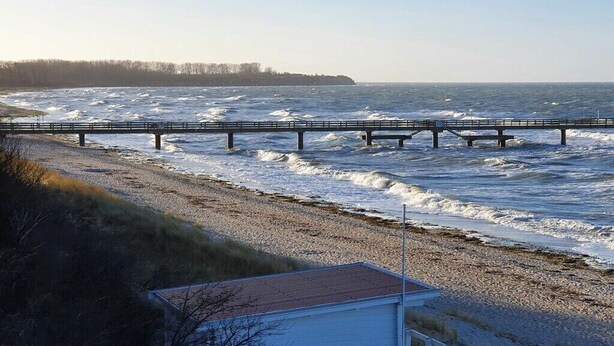 A coastal scene with a bridge over the water, a beach with pebbles, and a small building with a red roof. - Holiday Home by the Baltic Sea with Terrace (Ostseebad Rerik)