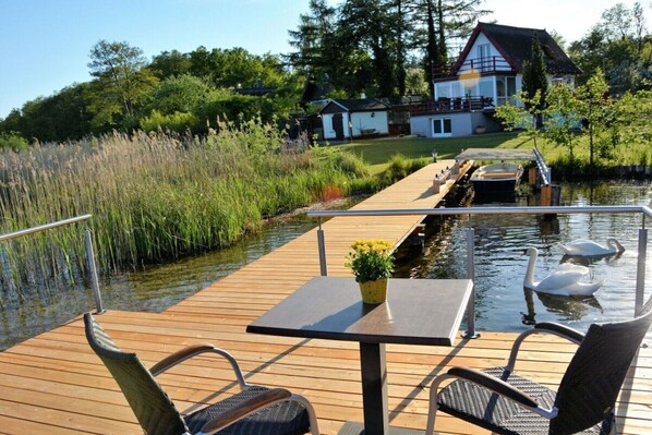Una terraza de madera con una mesa y sillas da a un cuerpo de agua tranquilo, con una casa al fondo.
