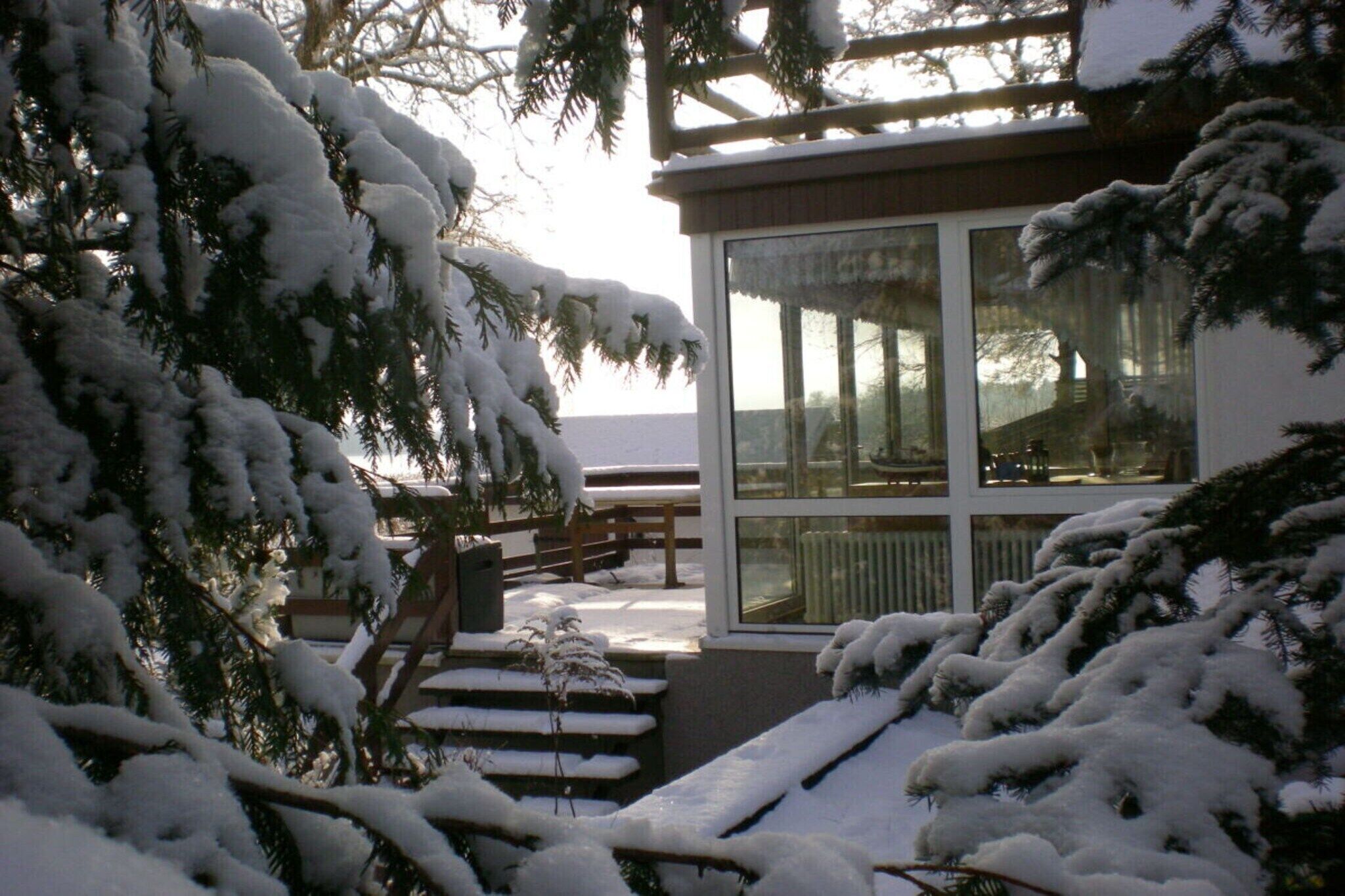 A snow-covered outdoor area with a wooden bench and a table, a building with large windows, and snow-laden trees.