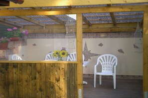 A covered outdoor seating area with a wooden table and chairs, a flower vase, and decorative wall elements. - Chalet by Lake with Sauna and Hot Tub (Krakow am See)