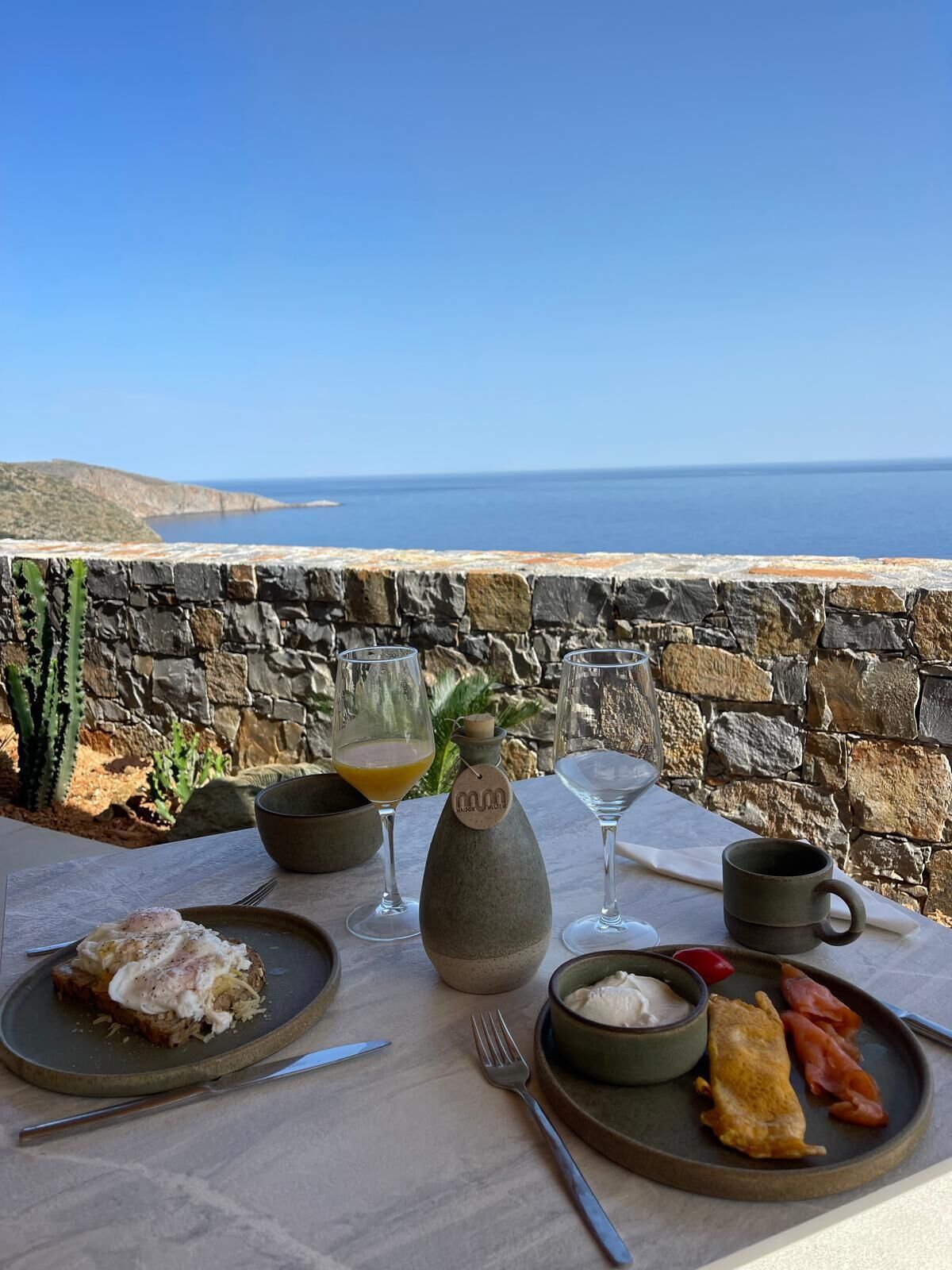 A table set with breakfast items including toast, eggs, and smoked salmon, with a view of the sea and a stone wall in the background.