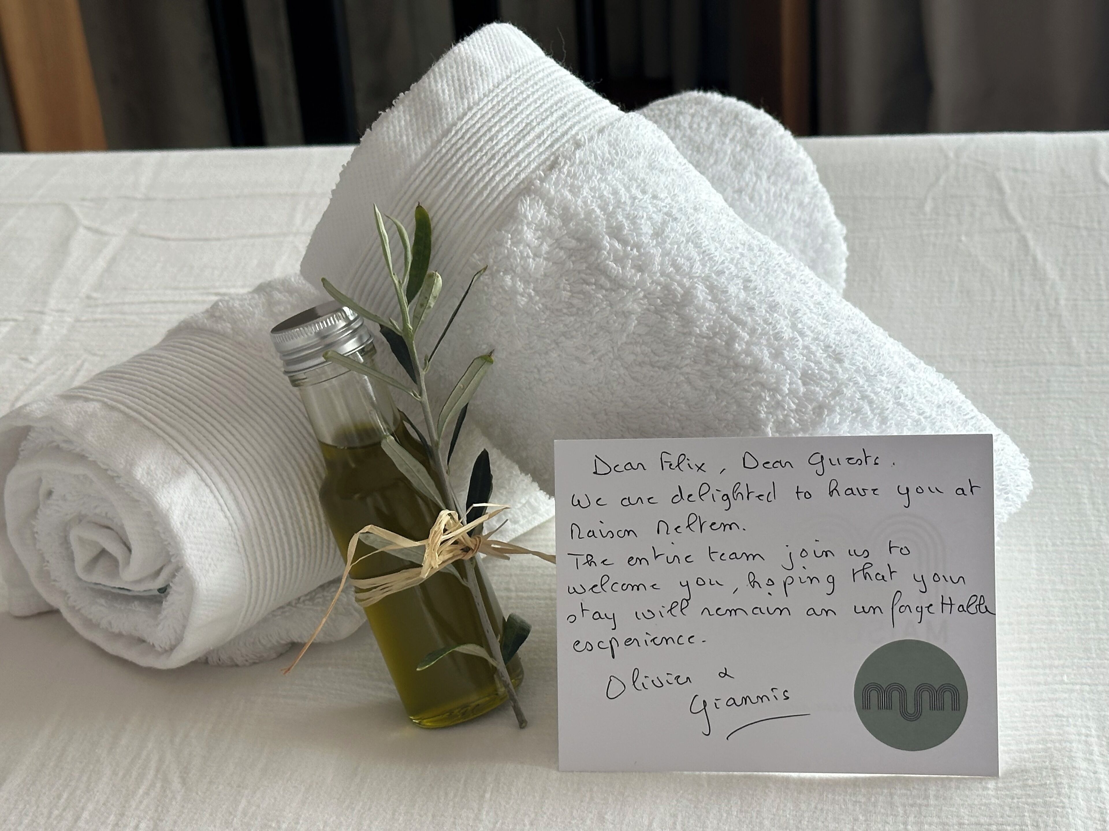 A rolled white towel, a small bottle with a plant, and a welcome card on a textured white surface.