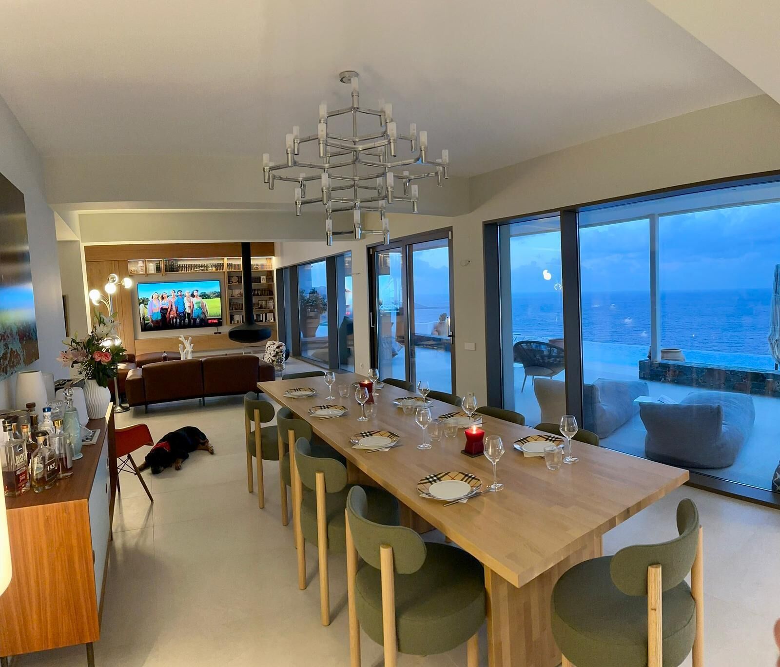 A modern dining area with a long table set for six, large windows offering a view of the sea, and a kitchen in the background.
